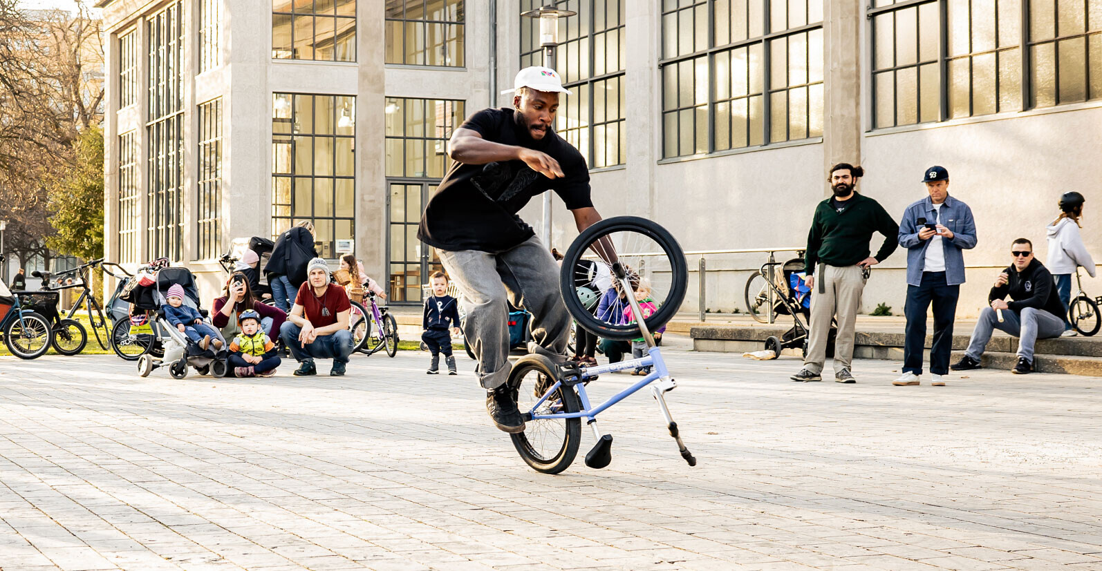 Omari Cato performing a flatland BMX trick in front of an audience in an urban square.