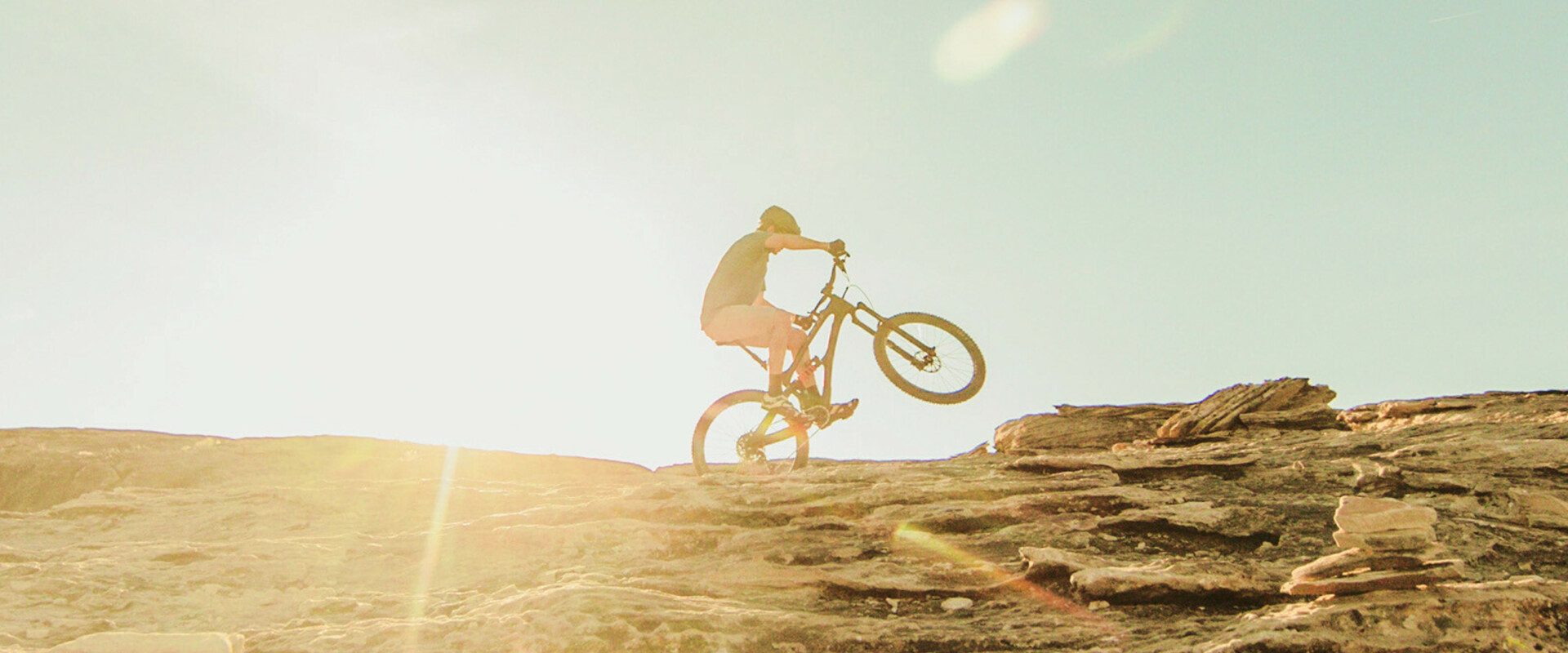 Mountain biker riding uphill on a rocky terrain during sunset, with sun rays visible in the background.