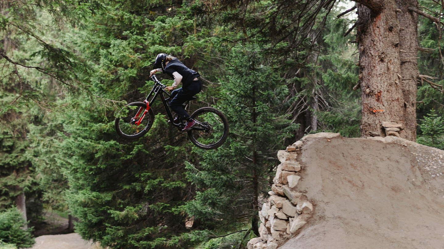 Un cycliste de montagne saute d'une rampe avec son vélo dans une forêt dense. Le cycliste porte un casque et des vêtements sombres, volant au-dessus d'un grand saut en terre.