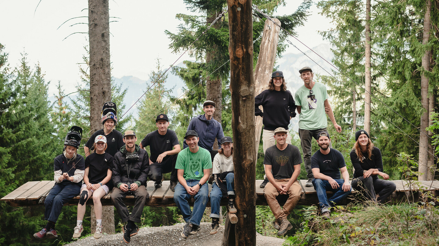 Un groupe de cyclistes de montagne et de membres de l'équipe posent ensemble sur une plateforme en bois dans la forêt. Les individus portent des vêtements sportifs et des casques, souriant à la caméra.