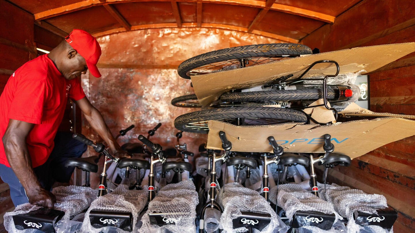 Un homme en chemise et casquette rouge charge des vélos dans une camionnette de livraison. Les vélos sont partiellement emballés et empilés. L'intérieur du véhicule a une teinte rouillée.