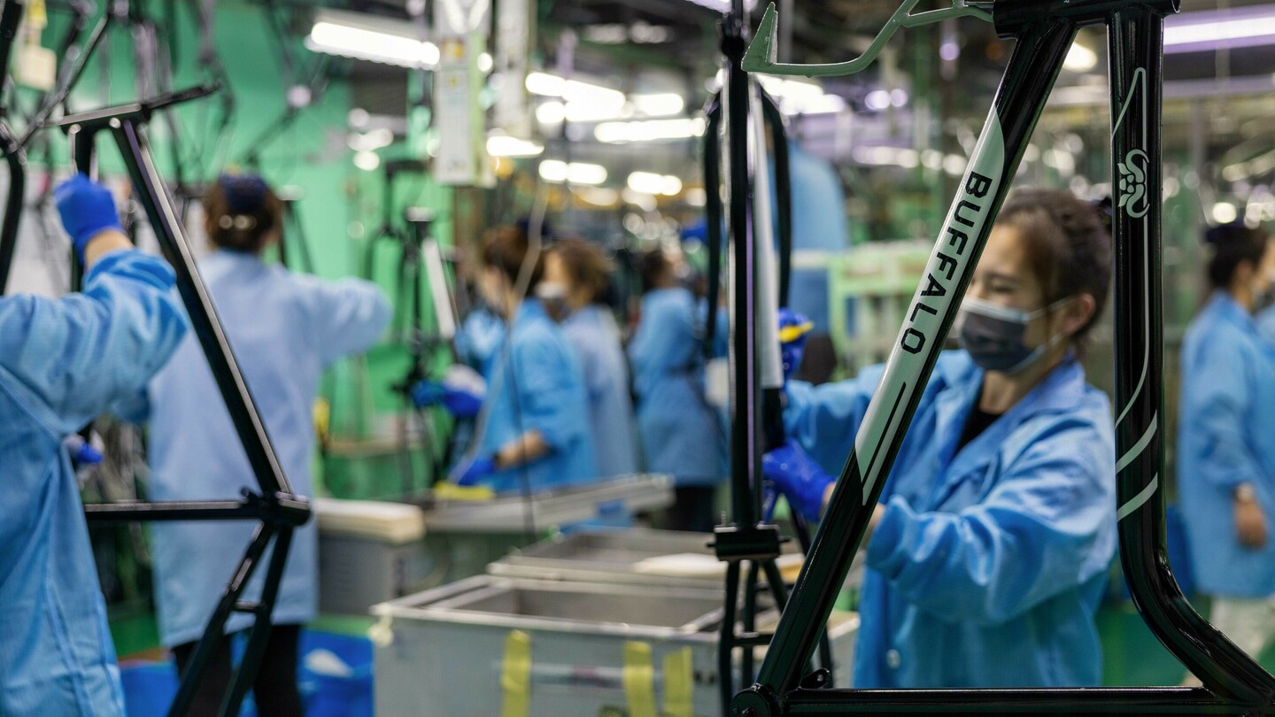 Atelier de production avec plusieurs personnes en blouses bleues et gants assemblant des cadres de vélo. L'un des cadres porte le logo “BUFFALO”. L'environnement de travail est industriel avec de nombreux outils et éclairages.