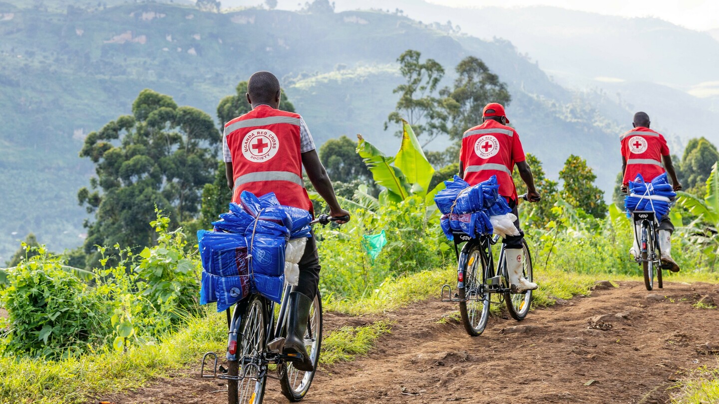 Three men in red vests with the Uganda Red Cross Society logo riding bicycles loaded with blue packages on a rural path through green landscape.