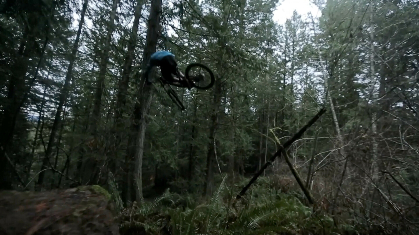 A mountain biker mid-air during a jump in a dense forest area. The rider is wearing dark clothing, and the forest is filled with tall trees and thick undergrowth.