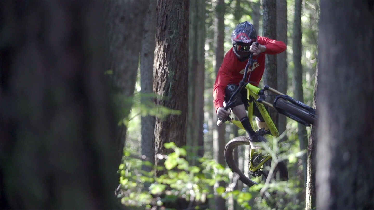 Mountain biker wearing a helmet and red shirt performing a jump in a dense forest, with the bike tilted sideways.