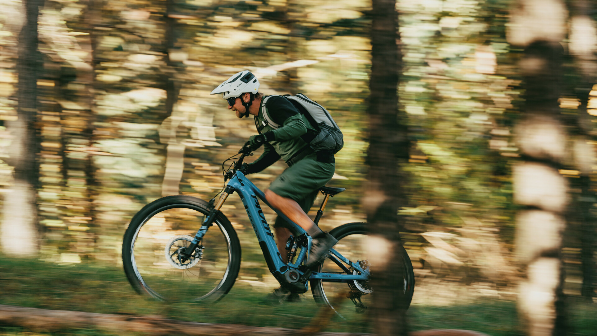 Mountain biker wearing a helmet and backpack riding through a forest at high speed on a blue bike.